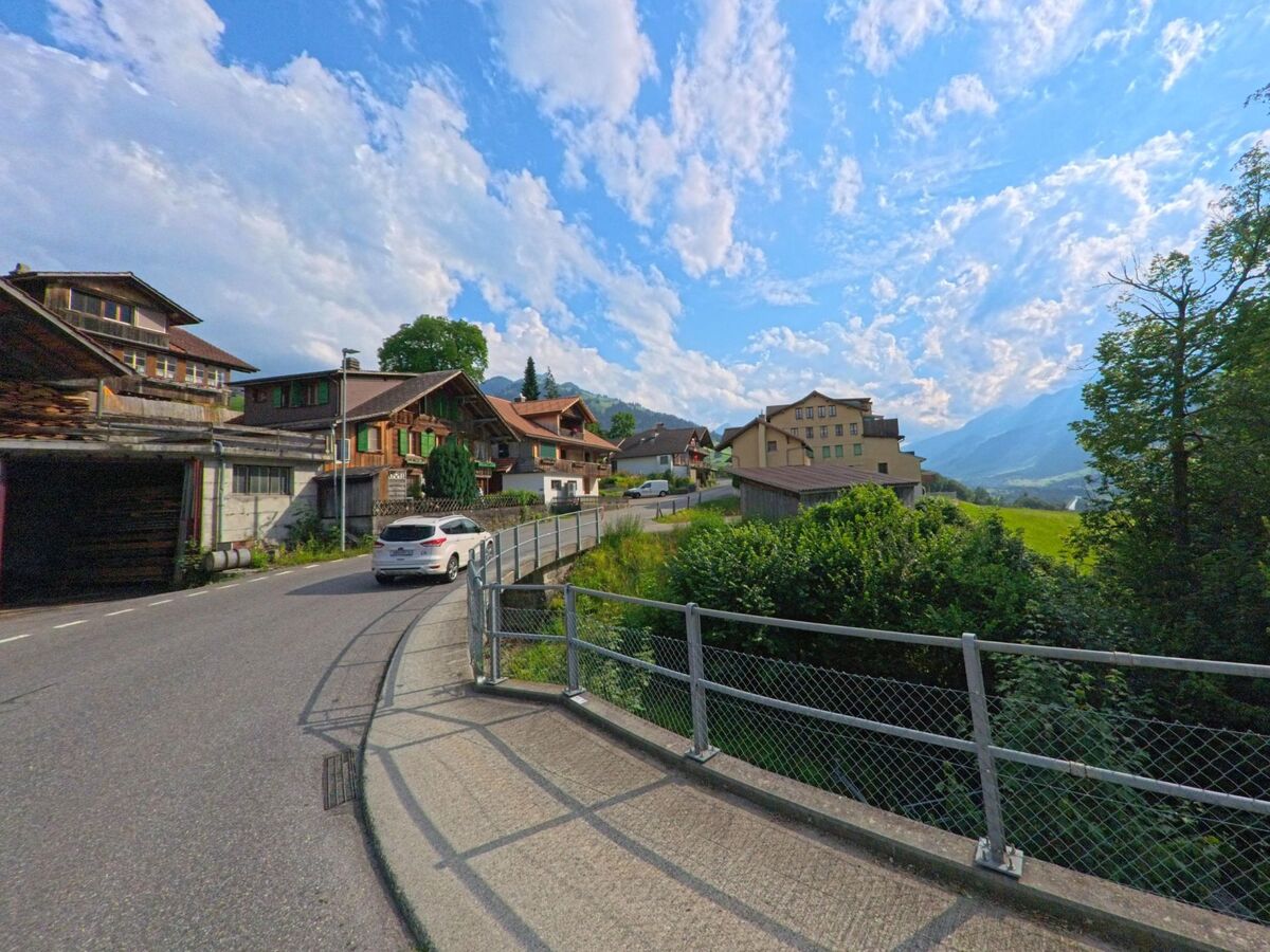 In Reichenbach im Kandertal (3722, Kanton Bern) windet sich die Strasse entlang traditioneller Holzhäuser und eines grösseren Gebäudes mit Blick auf das sonnige Tal und die Berge.