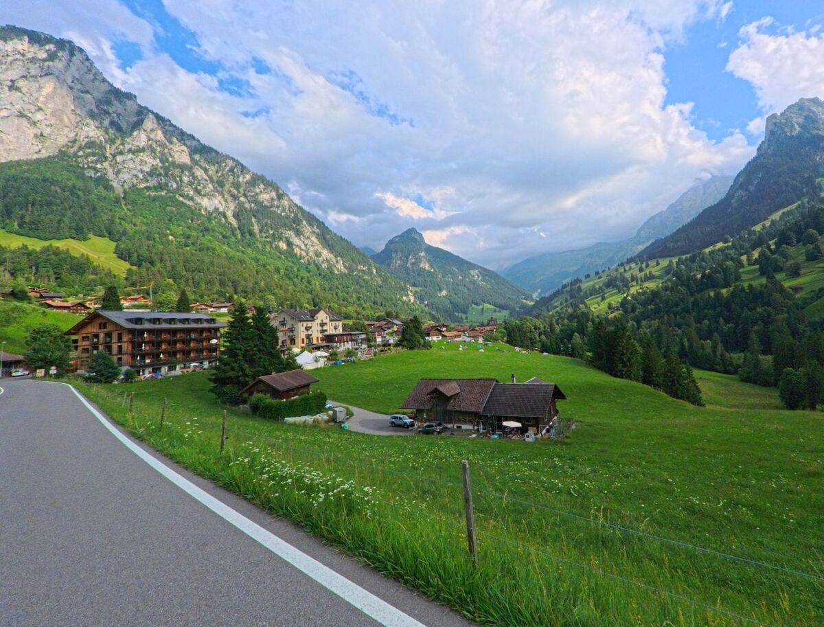 Entlang einer Strasse in Reichenbach im Kandertal (3723, Kanton Bern) erstreckt sich der Blick über verstreute Häuser, grüne Alpwiesen und steile Felswände in einem von Bergen eingerahmten Tal.