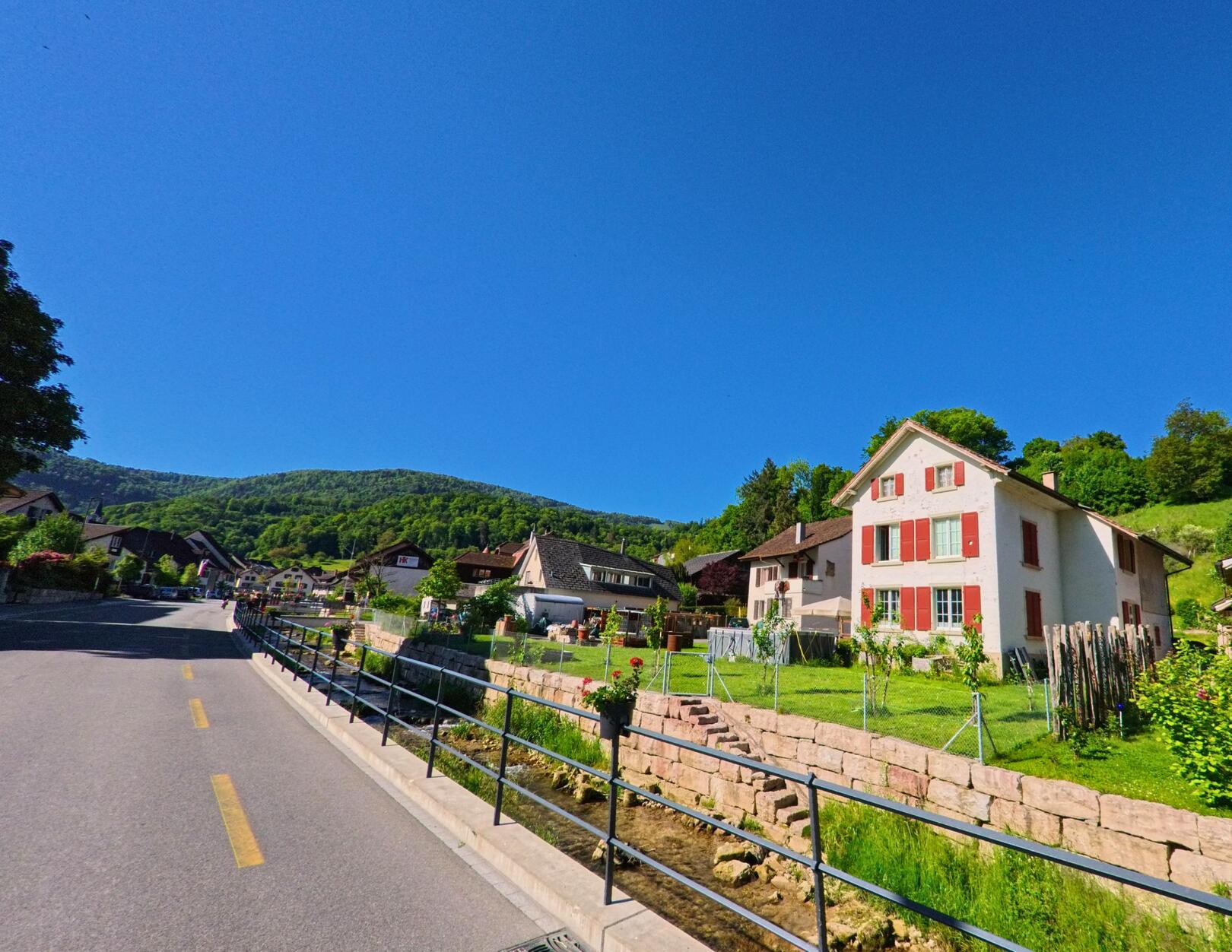 Entlang eines kleinen Bachs erstreckt sich in Reigoldswil (4418, Kanton Basel-Landschaft) eine ruhige Strasse mit gepflegten Wohnhaeusern und Blick auf bewaldete Huegel.