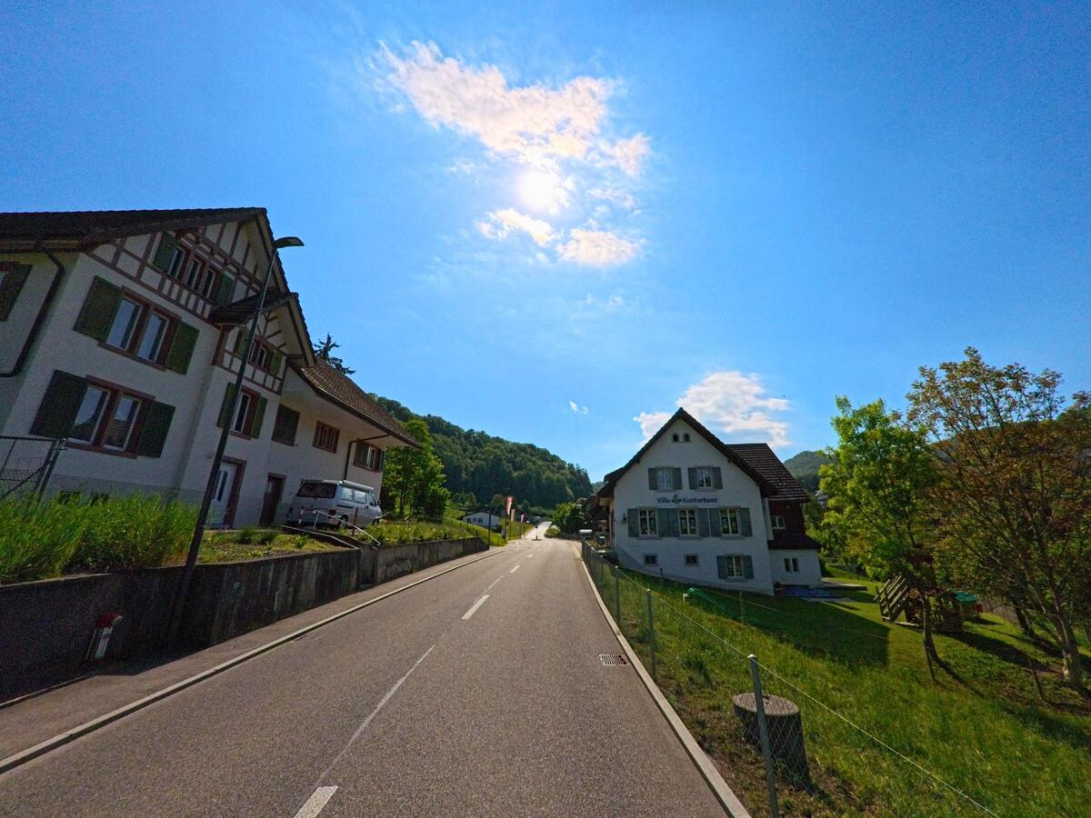 In Reigoldswil (4418, Kanton Basel-Landschaft) fuehrt eine leicht ansteigende Strasse zwischen traditionellen Wohnhaeusern und der Villa Kunterbunt vorbei in Richtung bewaldeter Hang.