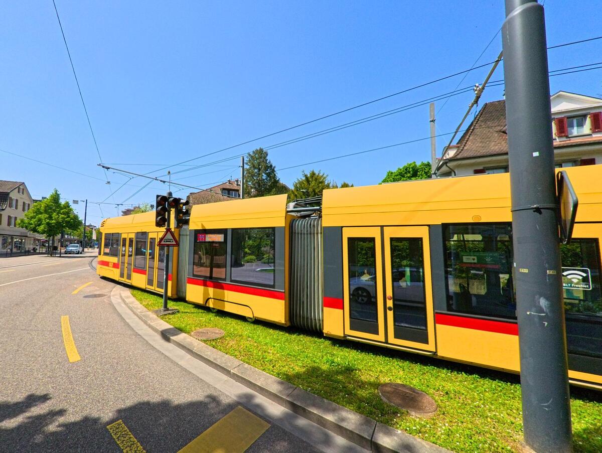 Eine gelbe Tram der BLT fährt durch Reinach (4153, Kanton Basel-Landschaft) an einer Kurve entlang, flankiert von Verkehrsampeln und Radstreifen.