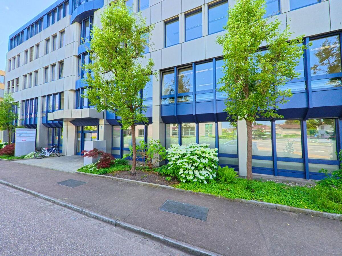 In Reinach (4153, Kanton Basel-Landschaft) steht ein modernes Bürogebäude mit blauen Fensterrahmen und gepflegtem Vorgarten entlang eines ruhigen Trottoirs.