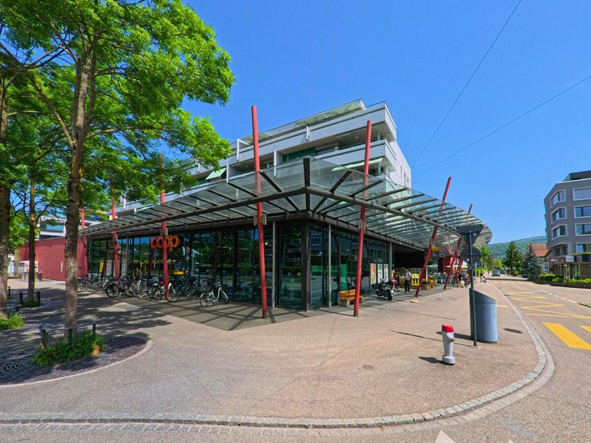Das moderne Coop-Gebäude mit markanter Glasfassade und roter Stahlkonstruktion in Reinach (4153, Kanton Basel-Landschaft) liegt an einer verkehrsberuhigten Strasse mit zahlreichen Veloständern.