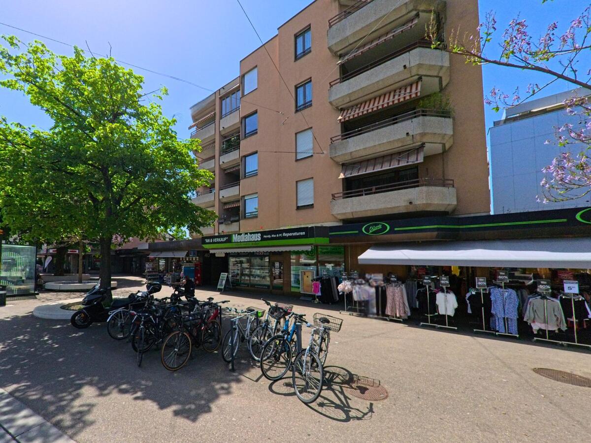 Im Zentrum von Reinach (PLZ 4153, Kanton Basel-Landschaft) befinden sich unter einem Wohnhaus mehrere Geschäfte mit Auslagen im Freien, davor steht ein Veloständer mit zahlreichen Fahrrädern.