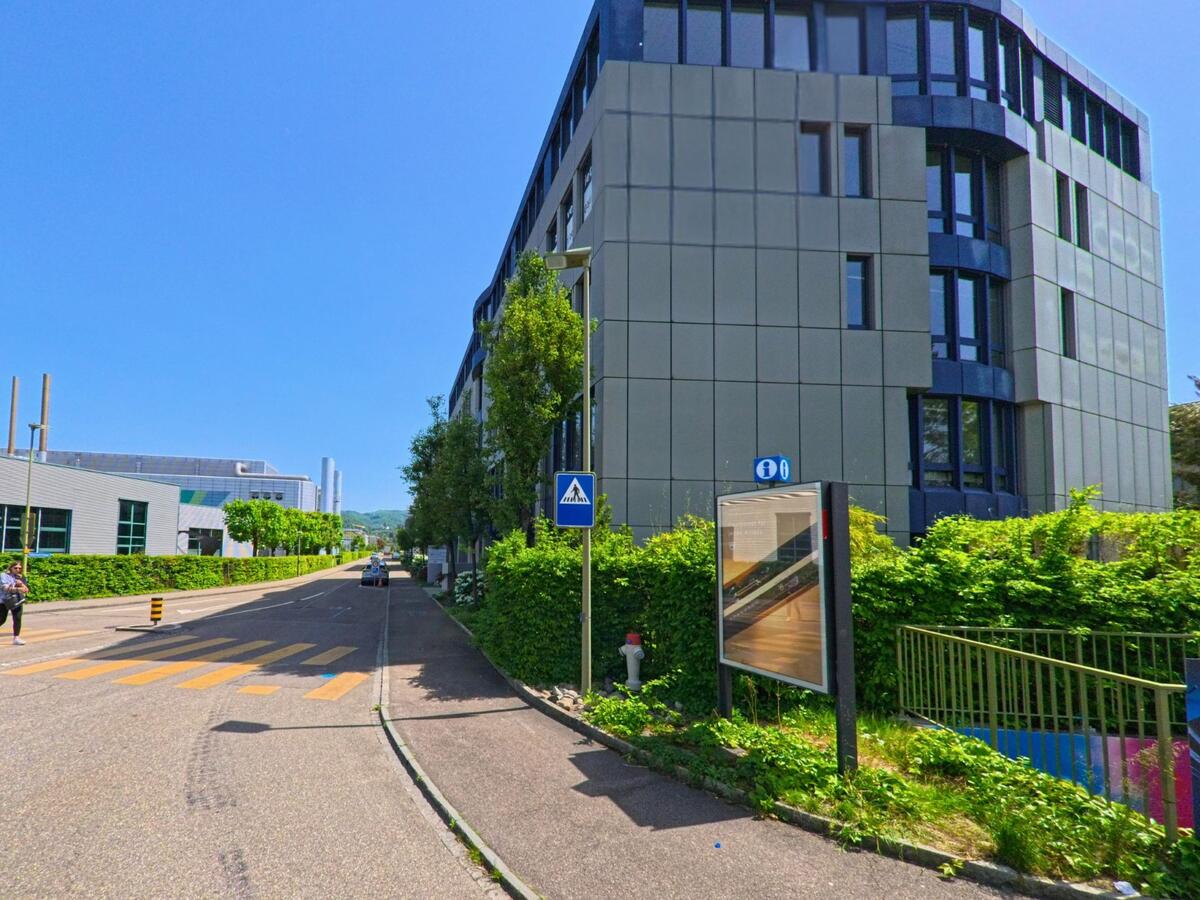 In Reinach (PLZ 4153, Kanton Basel-Landschaft) verläuft eine ruhige Strasse mit Fussgängerstreifen zwischen modernen Büro- und Industriegebäuden unter blauem Himmel.