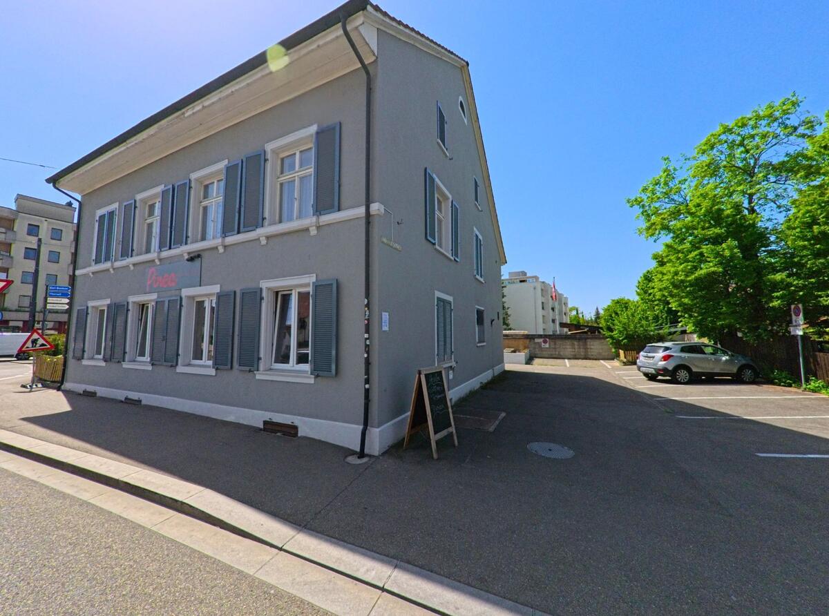 An einer Strassenecke in Reinach (4153, Kanton Basel-Landschaft) steht ein grau gestrichenes Gebäude mit Fensterläden, in dem sich ein Restaurant mit kleiner Tafel vor dem Eingang befindet.