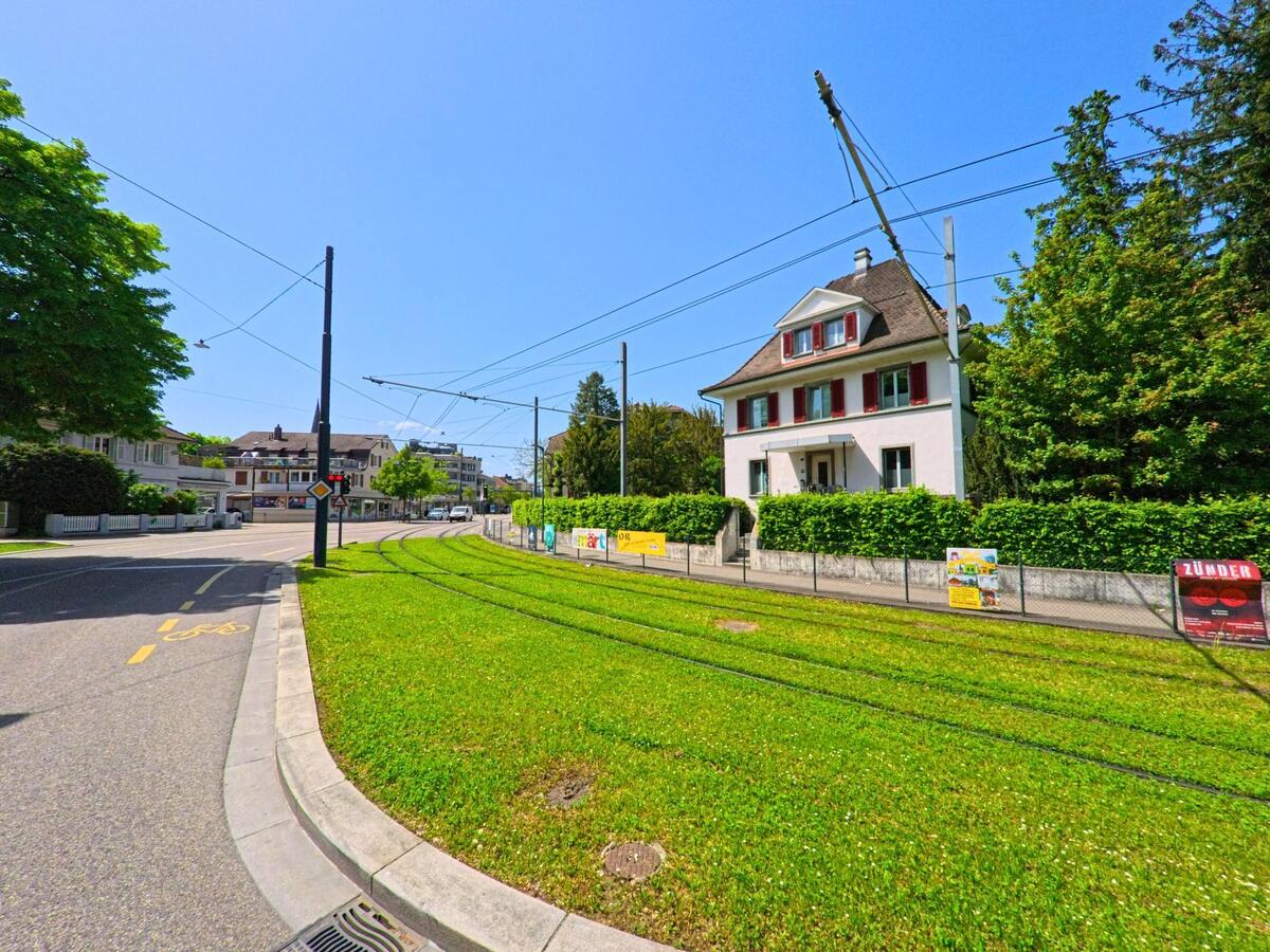 Eine Tramlinie mit begrüntem Gleisbett verläuft in Reinach (4153, Kanton Basel-Landschaft) an einem gepflegten Wohnhaus mit roten Fensterläden vorbei.