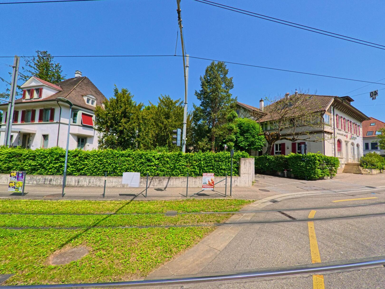 In Reinach (PLZ 4153, Kanton Basel-Landschaft) stehen entlang der Tramlinie zwei historische Wohnhäuser mit roten Fensterläden und dichtem Grünbewuchs im Vordergrund.