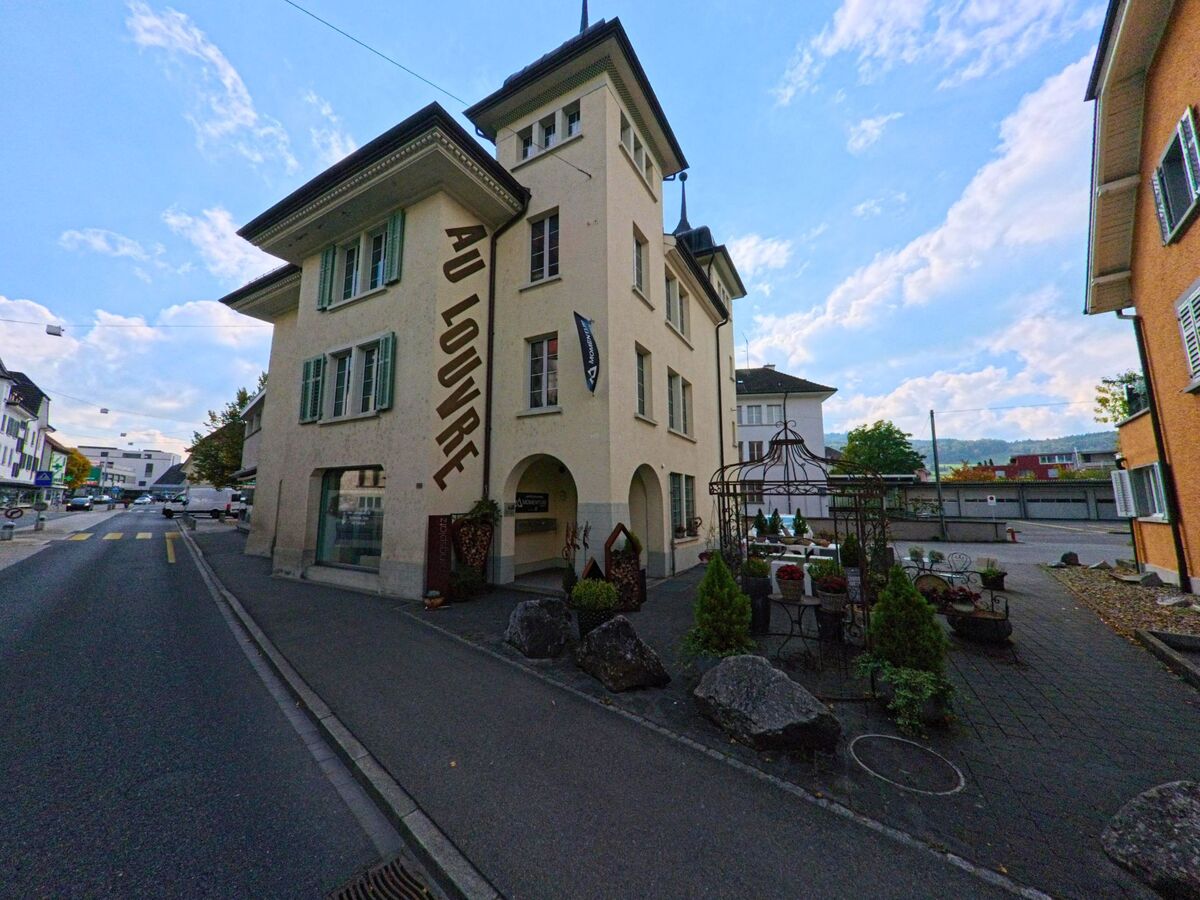 Das Gebäude des Restaurants «Au Louvre» in Reinach (5734, Kanton Aargau) steht an einer schmalen Strasse mit kleiner Gartenfläche und dekorierten Pflanzen.