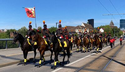 In Gunzwil (6222, Kanton Luzern) marschieren Reiter in Uniformen mit Pferden und Musikinstrumenten entlang einer Strasse, waehrend sie eine Fahne tragen und von Zuschauern bestaunt werden.