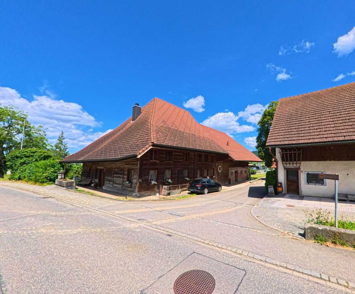 In Reitnau (5056, Kanton Aargau) stehen an einer Strassenecke zwei traditionelle Bauernhäuser mit markanten Walmdächern und Holzfassaden.