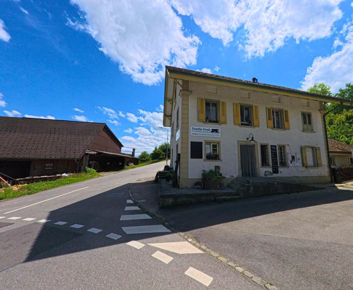 In Reitnau (5056, Kanton Aargau) steht ein Bauernhaus mit gelben Fensterläden an einer Weggabelung neben einem Wirtschaftsgebäude aus Holz.