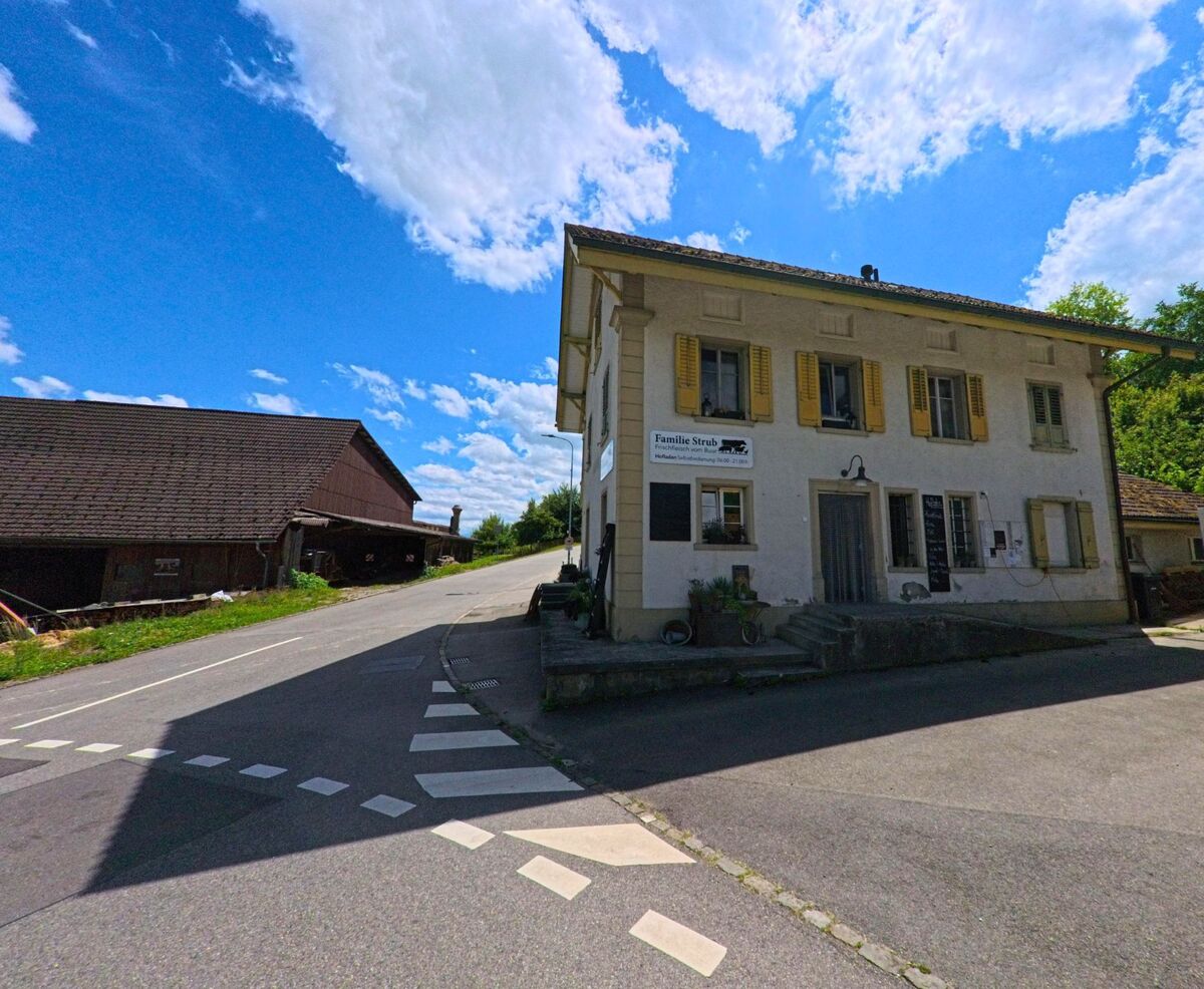 In Reitnau (5056, Kanton Aargau) steht ein Bauernhaus mit gelben Fensterläden an einer Weggabelung neben einem Wirtschaftsgebäude aus Holz.