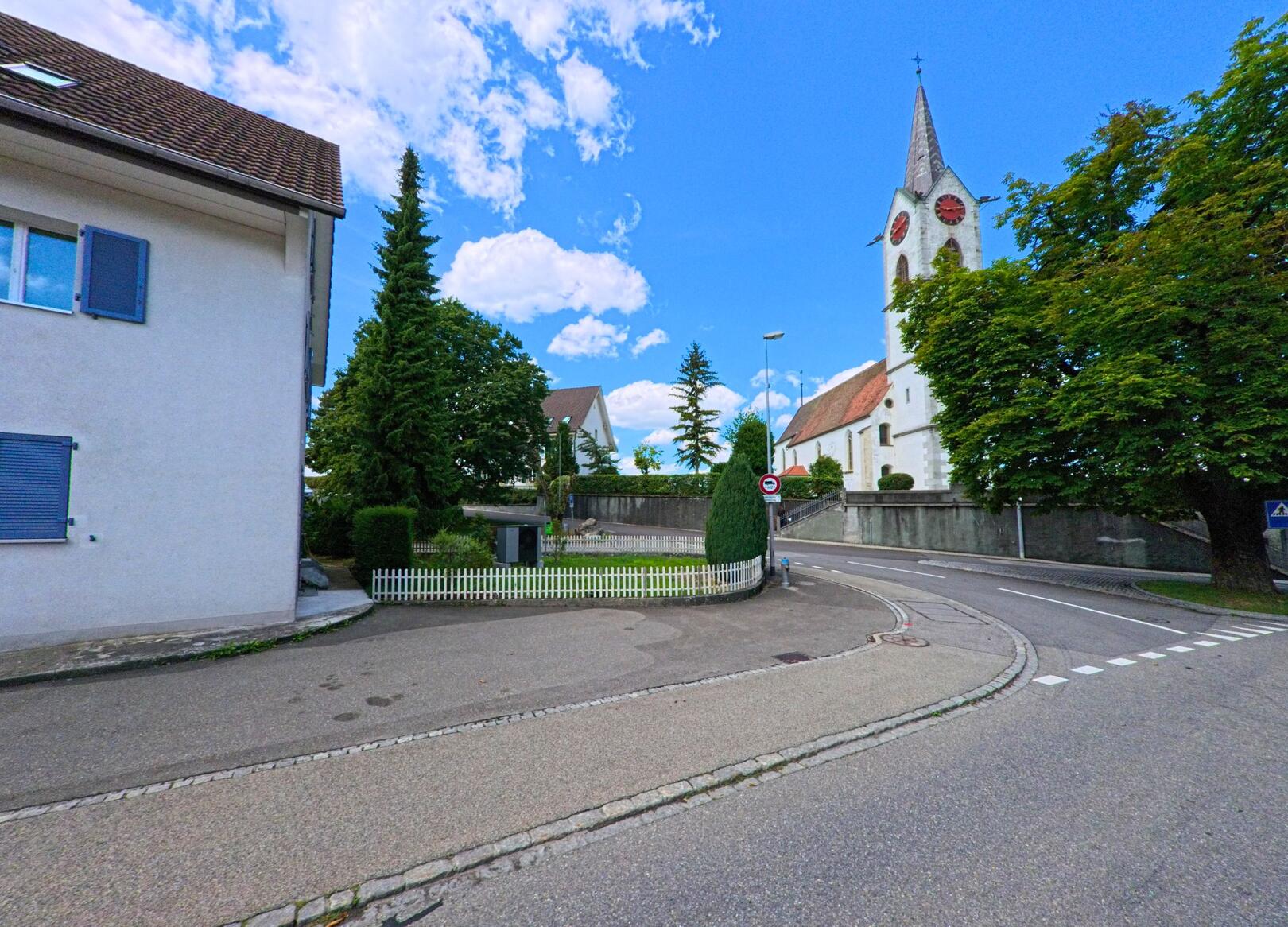 Die reformierte Kirche von Reitnau (5057, Kanton Aargau) mit ihrem markanten Turm steht oberhalb einer Kurve im Zentrum des Dorfes, umgeben von Bäumen und gepflegten Anlagen.
