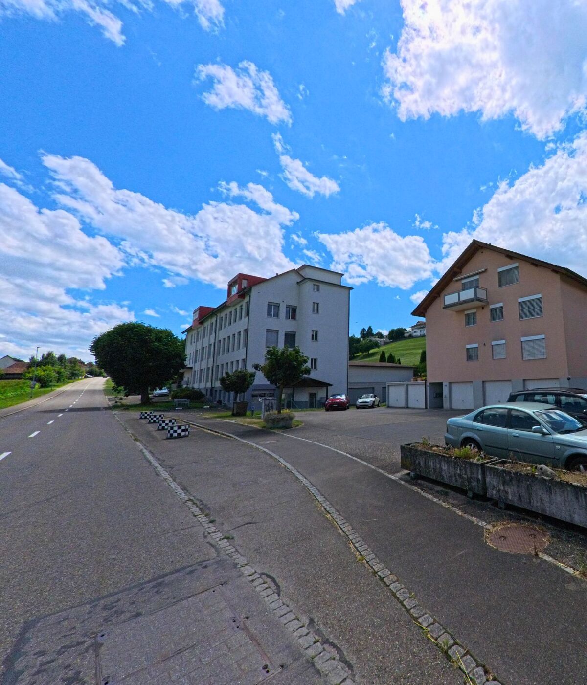 Entlang der Hauptstrasse von Reitnau (5057, Kanton Aargau) stehen mehrstöckige Wohngebäude mit Parkplätzen und Blick auf die angrenzenden Hügel.