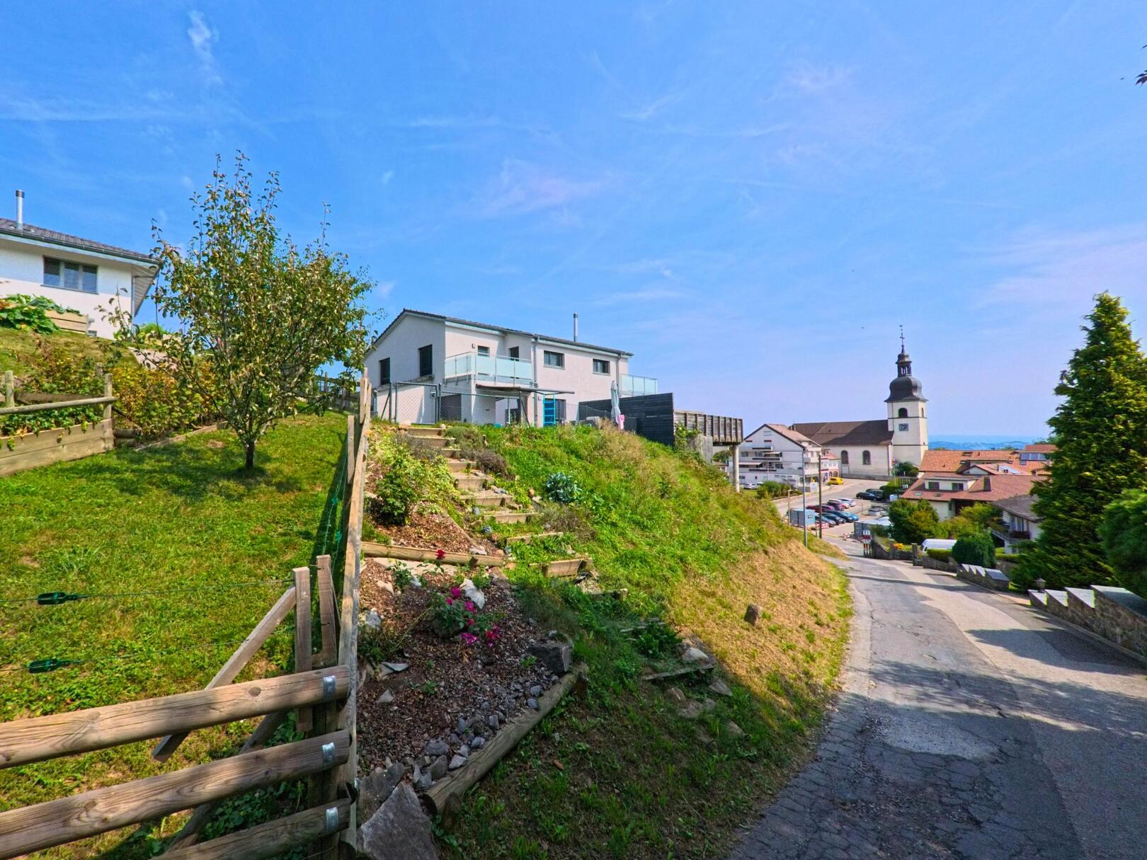 Ein Blick auf den Dorfkern von Remaufens (1617, Kanton Freiburg) zeigt die Kirche im Hintergrund und einen Weg, der an Gärten und Haeusern vorbeiführt.