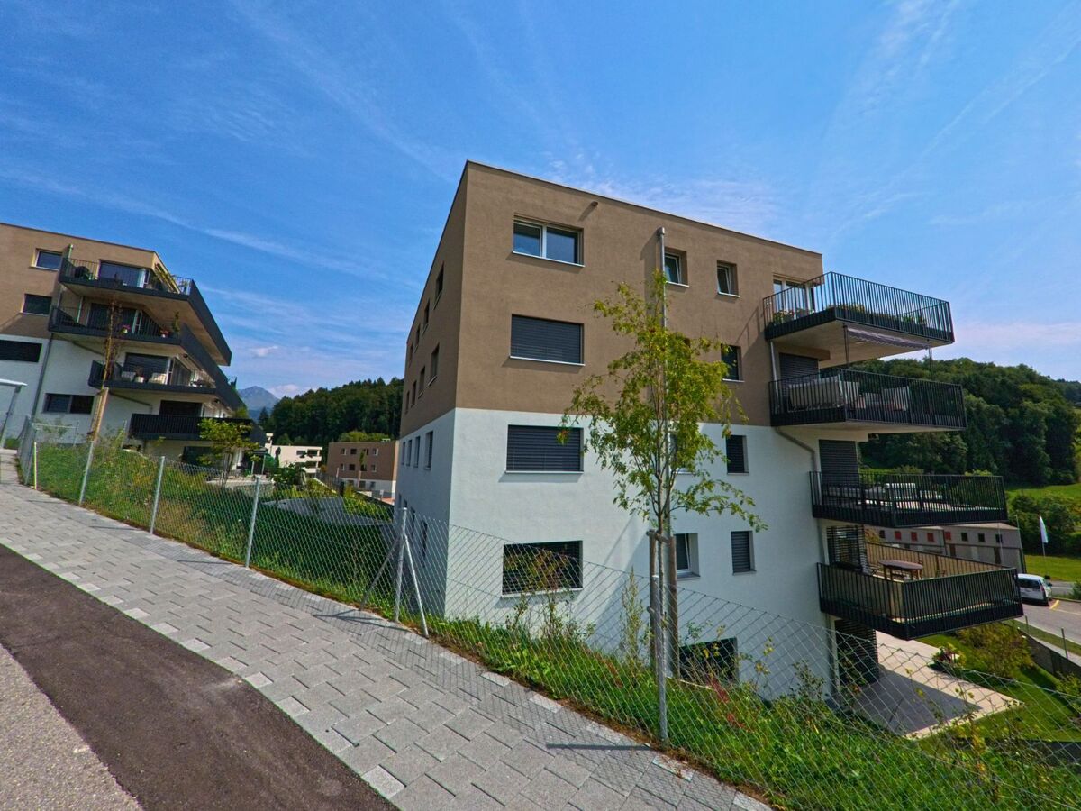 Ein Neubau mit mehreren Balkonen erhebt sich am Hang von Remaufens (1617, Kanton Freiburg), waehrend dahinter bewaldete Huegel die Landschaft praegen.