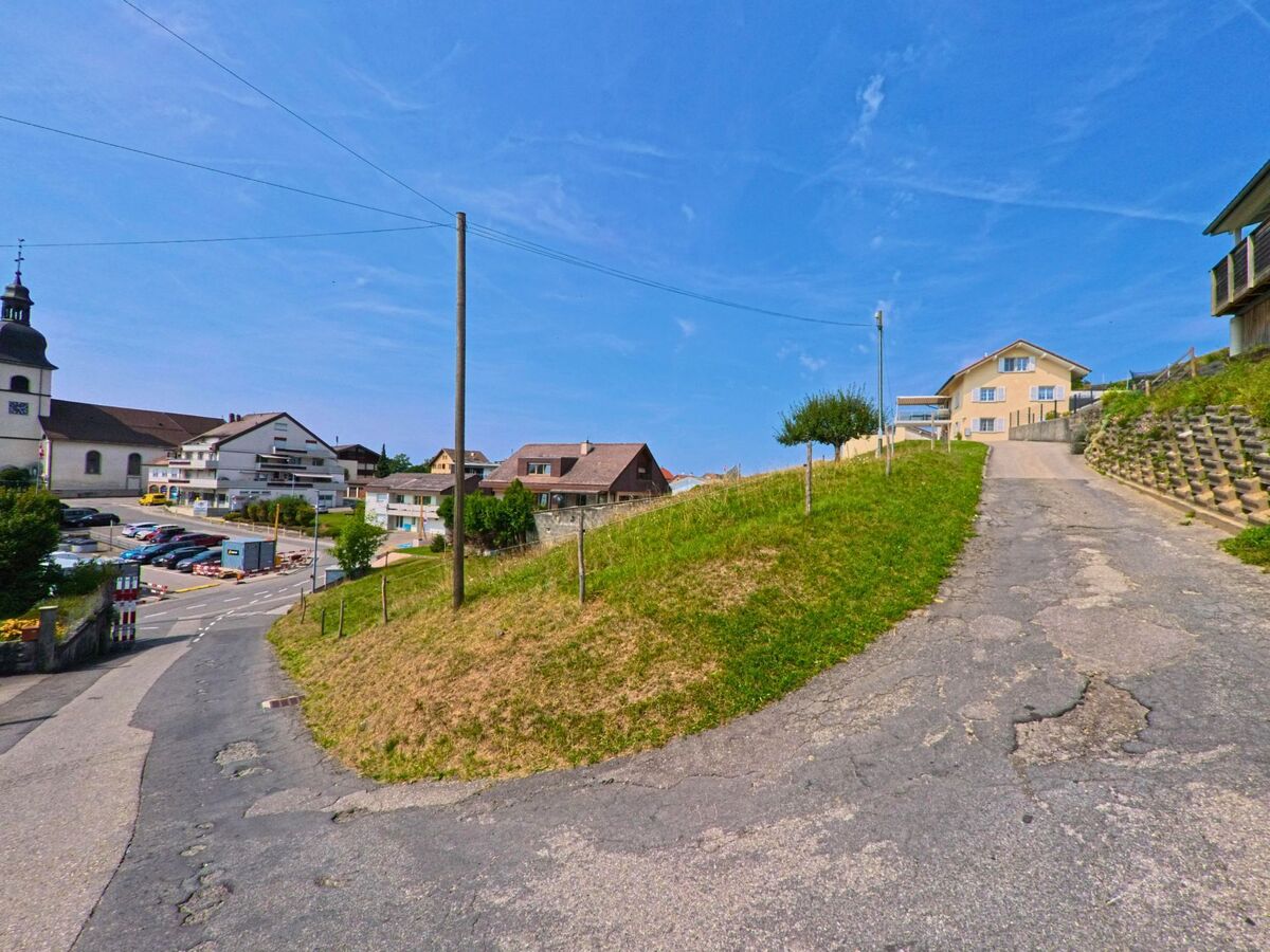 Eine steile Strasse fuehrt in Remaufens (1617, Kanton Freiburg) von Wohnhaeusern hinab in Richtung Dorfzentrum mit Kirche und Parkplaetzen.