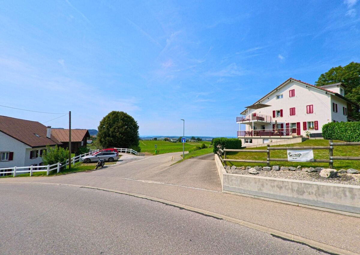 Am Ortsrand von Remaufens (1617, Kanton Freiburg) fuehrt eine Strasse an traditionellen und modernen Wohnhaeusern vorbei mit Blick auf die offene Landschaft.
