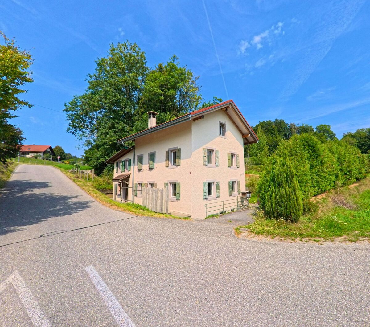 Ein helles Wohnhaus mit gruenen Fensterlaeden liegt an einer ansteigenden Strasse in Remaufens (1617, Kanton Freiburg) nahe eines bewaldeten Hangs.