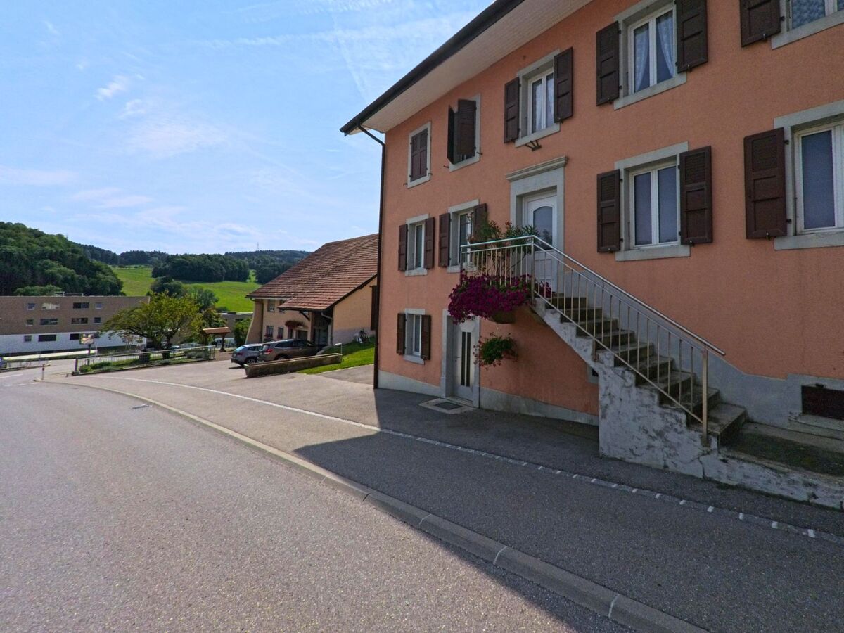 Ein lachsfarbenes Wohnhaus mit Aussentreppe und Blumenschmuck steht an einer Strasse in Remaufens (1617, Kanton Freiburg) mit Blick auf Wiesen und Huegel.