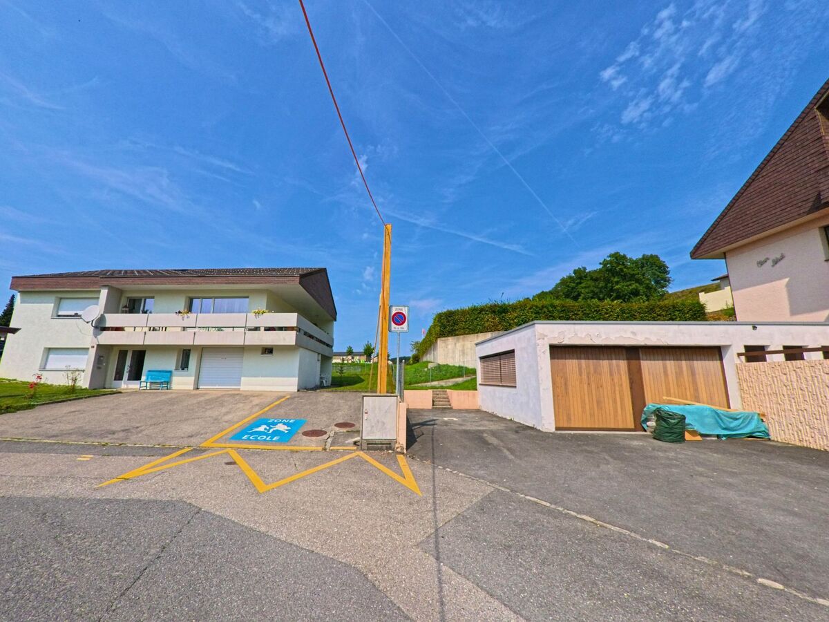 Ein Wohnhaus mit Balkonen und ein separates Garagengebaeude befinden sich an einer Strasse in Remaufens (1617, Kanton Freiburg) nahe einer markierten Schulzone.