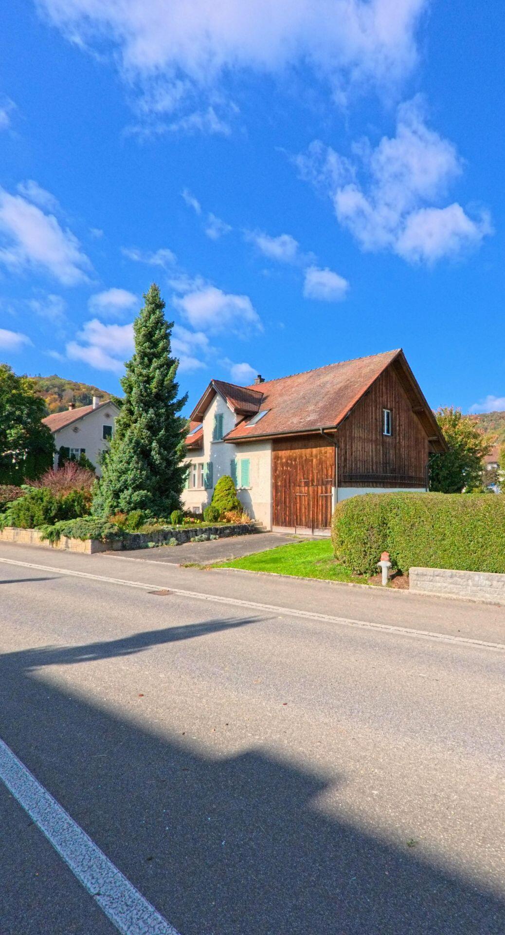 Ein Einfamilienhaus mit Holzfassade und grossem Nadelbaum steht an einer Nebenstrasse in Remigen (5236, Kanton Aargau) vor bewaldeten Hügeln.