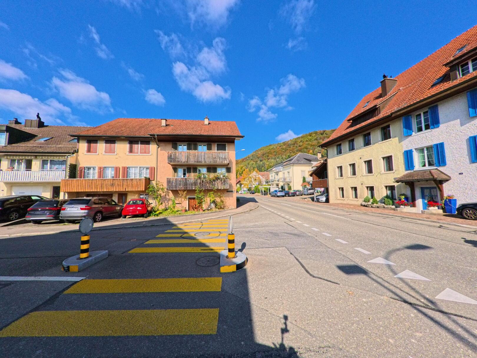 In Remigen (5236, Kanton Aargau) verläuft eine Hauptstrasse mit Fussgängerstreifen zwischen farbenfrohen Wohnhäusern unter einem klaren blauen Himmel.