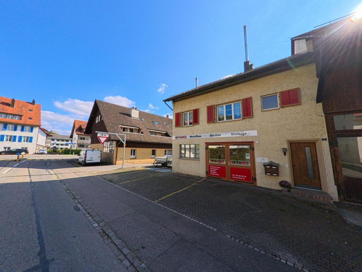 In Remigen (5236, Kanton Aargau) befindet sich ein Gewerbehaus mit roter Beschriftung an einer ruhigen Strasse, umgeben von Wohnhäusern mit Satteldächern.