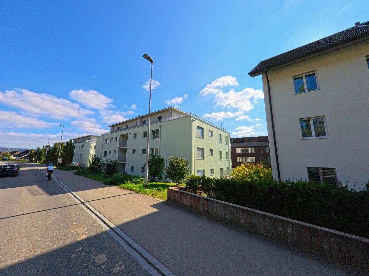 Mehrere moderne Mehrfamilienhäuser mit Balkonen stehen an einer sonnigen Strasse in Remigen (5236, Kanton Aargau), während ein Radfahrer vorbeifährt.