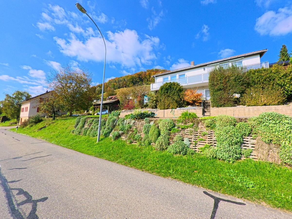 Ein Einfamilienhaus mit grossem Balkon und terrassiertem Vorgarten liegt an einer leicht ansteigenden Strasse in Remigen (5236, Kanton Aargau).