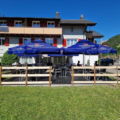 Vor einem Gebaeude mit roten Fensterlaeden in Unteriberg (8842, Kanton Schwyz) stehen auf einer Terrasse mehrere Tische unter blauen Sonnenschirmen.