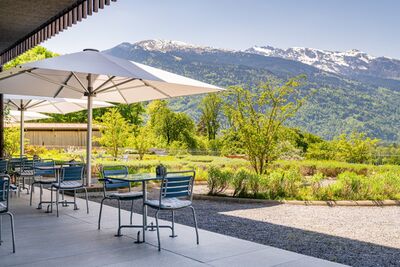 In Flaesch (7306, Kanton Graubuenden) stehen Tische mit Sonnenschirmen auf einer Terrasse, die einen weiten Blick auf gruen bewaldete Berghaenge bietet.