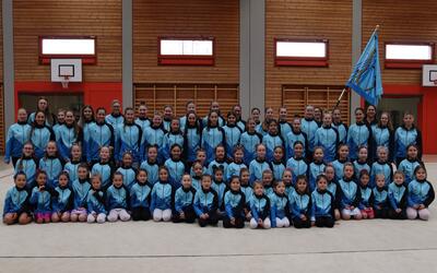 Die Mitglieder der RG/Gym Diepoldsau-Schmitter posieren in Diepoldsau (9444, Kanton St. Gallen) in blau-schwarzen Trainingsanzügen mit Vereinsfahne für ein Gruppenfoto in einer Sporthalle.