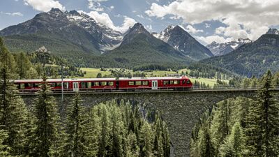 In Chur (7001, Kanton Graubuenden) ueberquert ein roter Zug der Rhaetischen Bahn eine steinerne Bruecke vor beeindruckender Alpenkulisse.