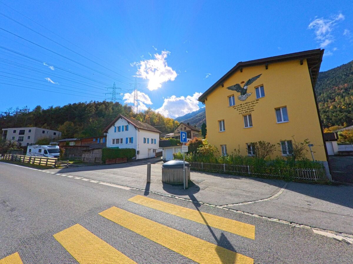 An einem Fussgängerstreifen in Rhäzüns (7403, Kanton Graubünden) stehen Wohnhäuser mit farbigen Fassaden, während sich dahinter bewaldete Berghänge unter klarem Himmel erheben.