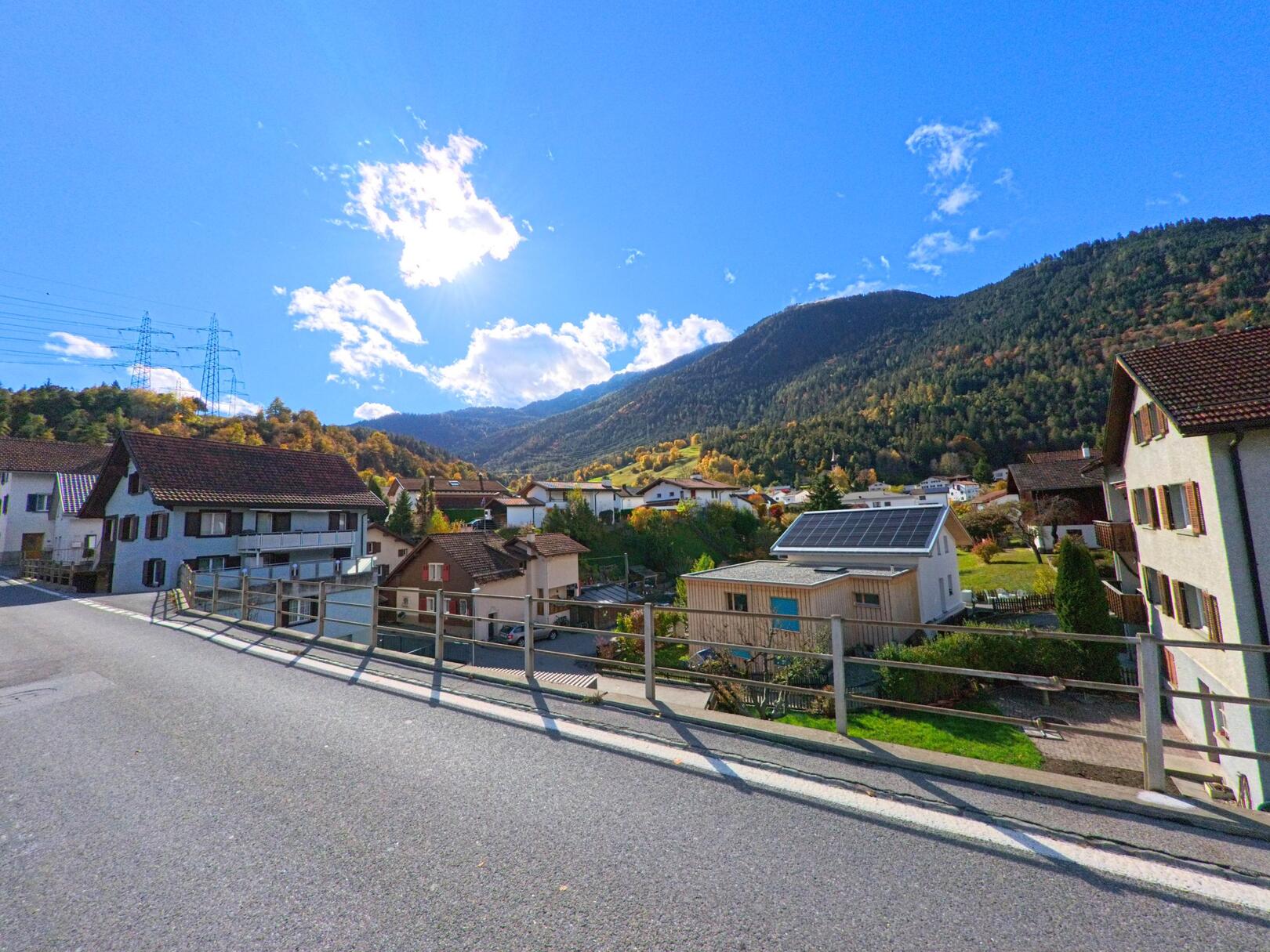 Von einer leicht erhöhten Strasse in Rhäzüns (7403, Kanton Graubünden) fällt der Blick auf Wohnhäuser und Gärten, die sich vor bewaldeten Berghängen im klaren Tageslicht ausbreiten.