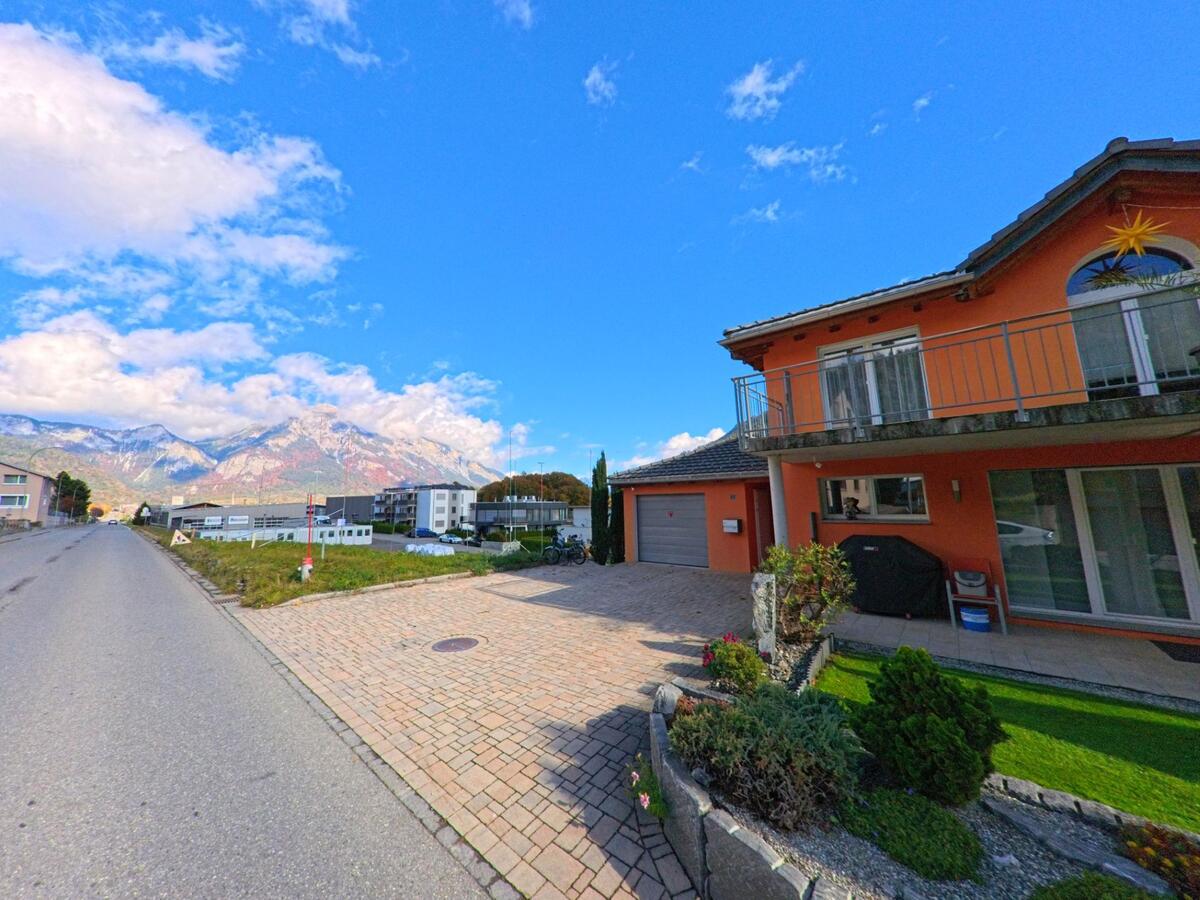 An einer ruhigen Strasse in Rhäzüns (7403, Kanton Graubünden) steht ein Wohnhaus mit gepflegtem Vorgarten, während sich im Hintergrund Gewerbebauten und markante Bergzüge zeigen.