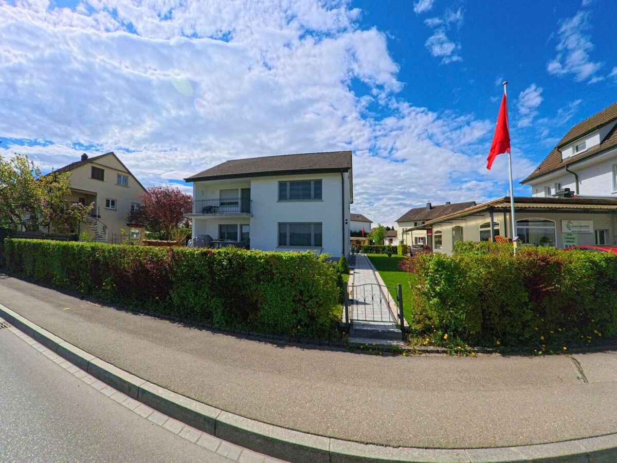 Eine gepflegte Wohnanlage mit Schweizer Fahne vor dem Eingang zeigt sich in Rheinau (8462, Kanton Zürich) unter einem teilweise bewölkten Himmel entlang einer ruhigen Quartierstrasse.