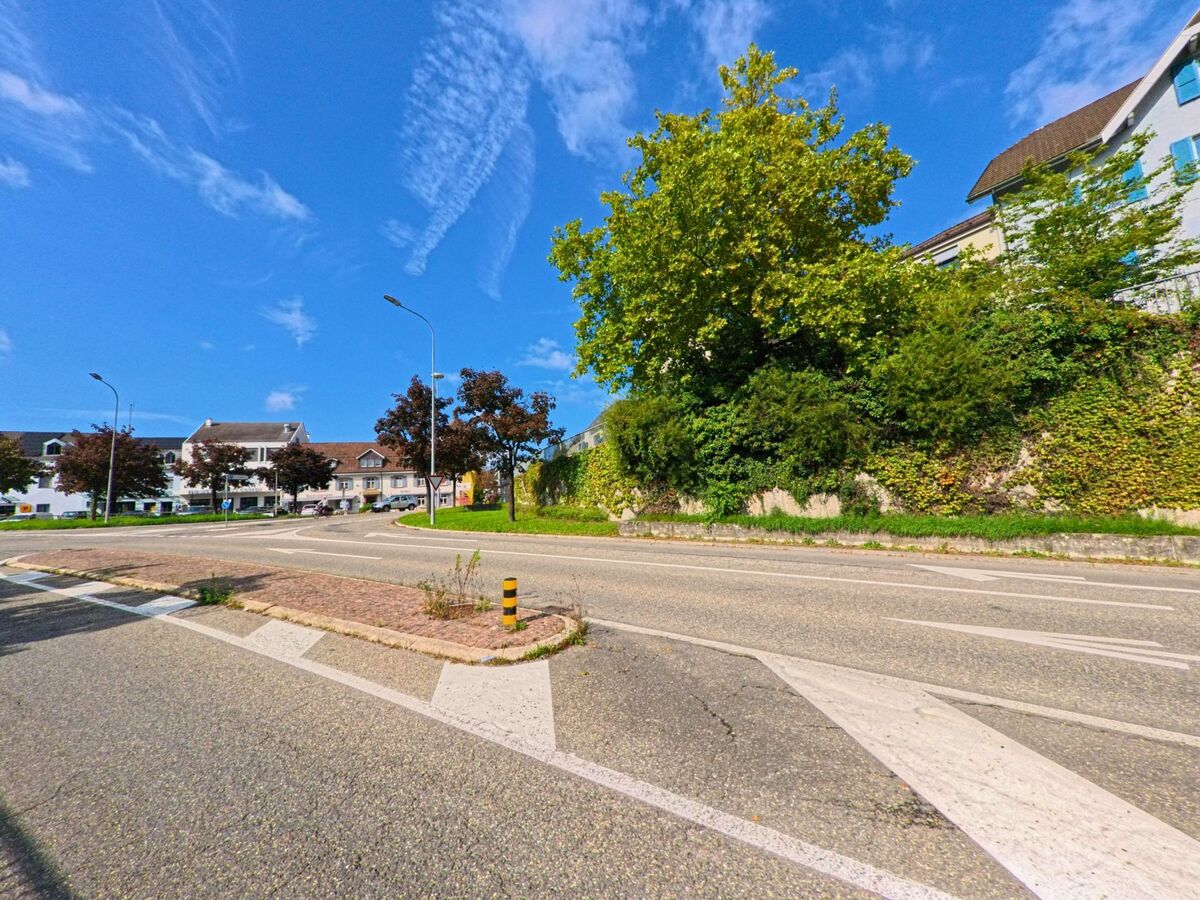 Eine Kreuzung in Rheinfelden (4310, Kanton Aargau) zeigt begruen­te Mauern und Wohnhaeuser unter einem klaren blauen Himmel.