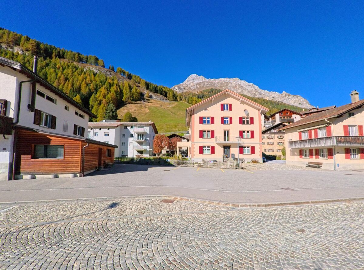 Zwischen gepflegten Wohnhäusern öffnet sich in Rheinwald (7435, Kanton Graubünden) ein kleiner Platz, der von gepflasterter Fahrbahn und alpiner Hanglandschaft umrahmt wird.