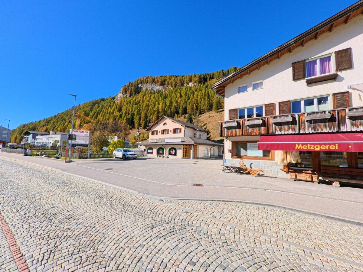 An einer gepflasterten Ortsstrasse zeigt sich in Rheinwald (7435, Kanton Graubünden) ein kleiner Dorfkern mit Geschäften und Wohnhäusern vor einem bewaldeten Berghang.