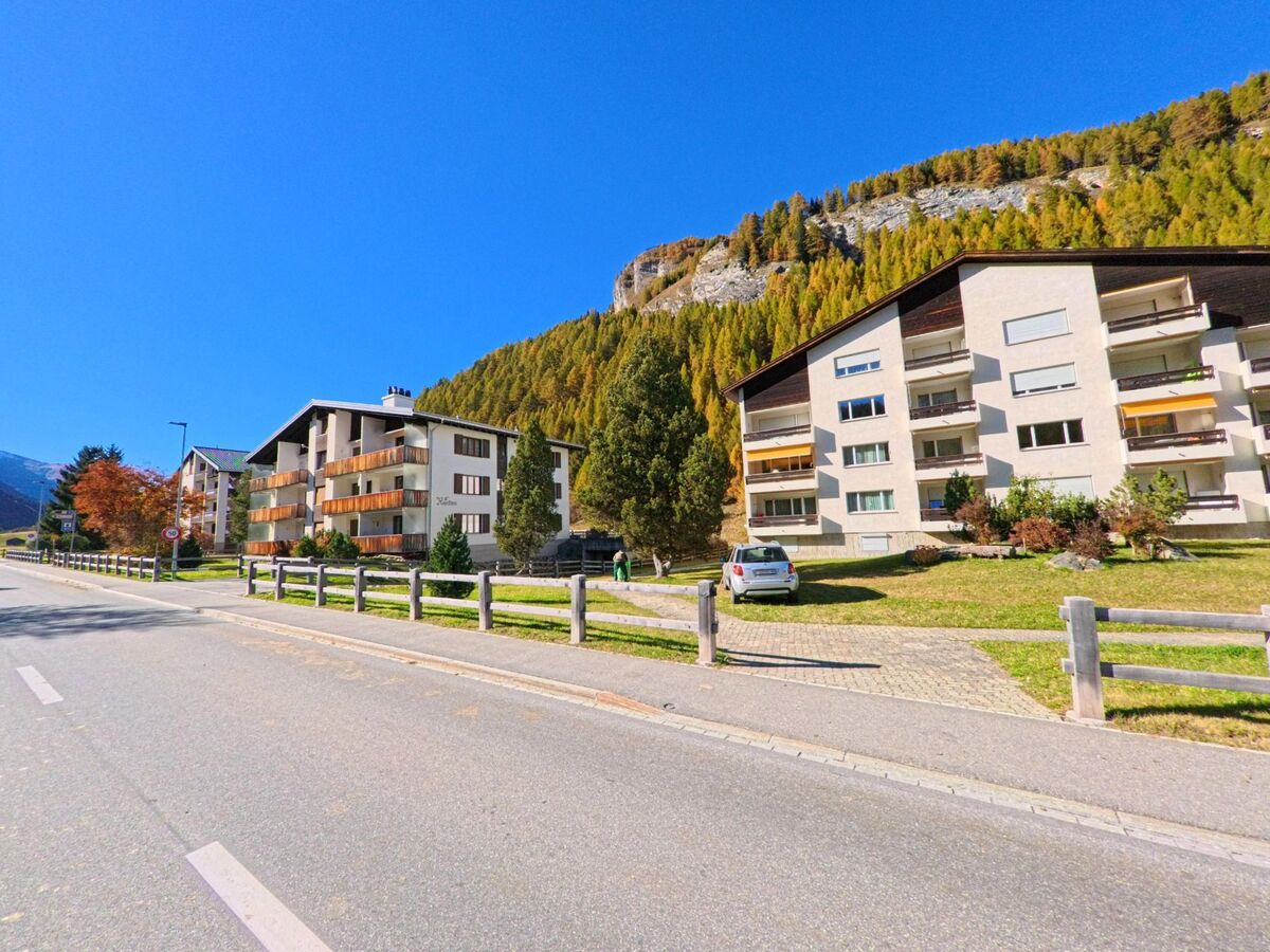 Am Rand einer breiten Talstrasse stehen in Rheinwald (7435, Kanton Graubünden) moderne Wohnhäuser mit Balkonen vor einem steil ansteigenden, herbstlich gefärbten Berghang.