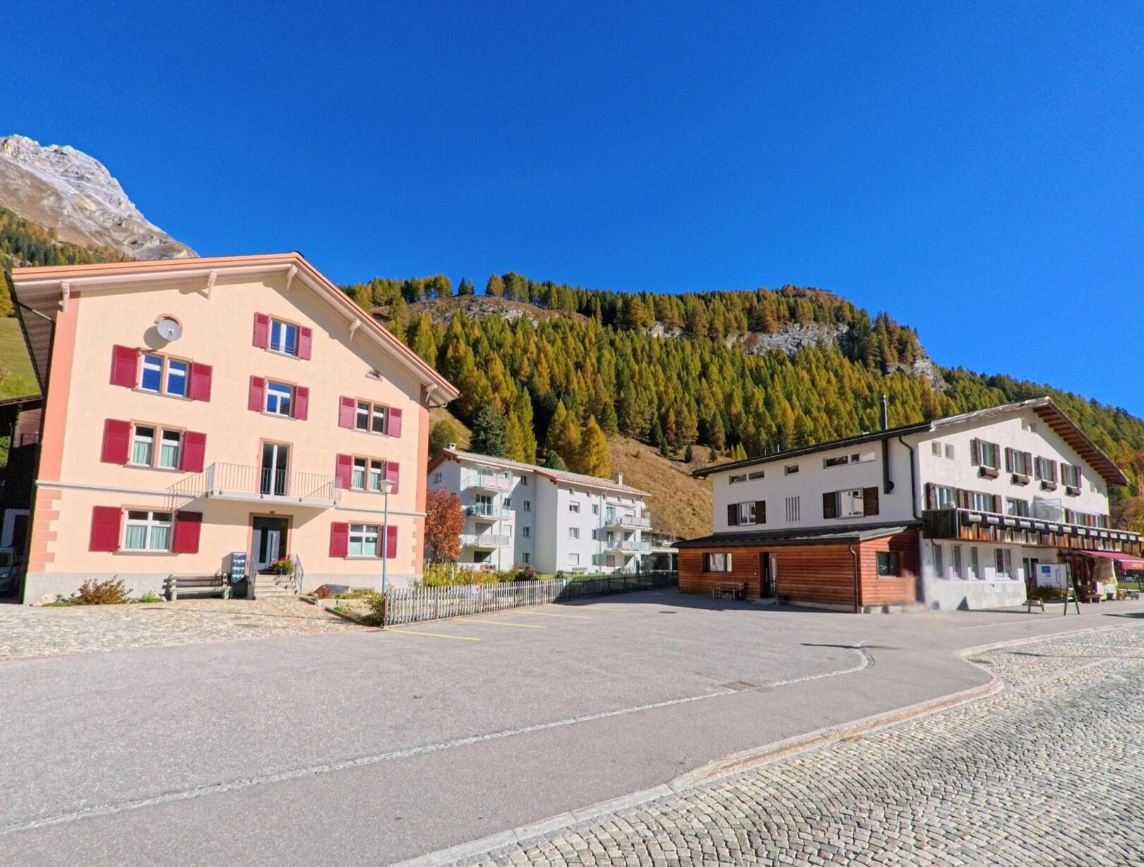 Vor bewaldeten Berghängen zeigt sich in Rheinwald (7435, Kanton Graubünden) ein Platz mit Wohnhäusern und einem Gasthaus entlang einer gepflasterten Strasse.