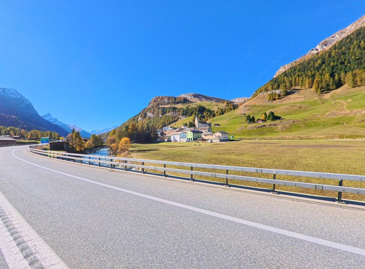 Von einer leicht geschwungenen Strasse aus öffnet sich in Rheinwald (7435, Kanton Graubünden) der Blick auf ein Dorf am Talboden mit Flusslauf und weitläufigen Wiesen.