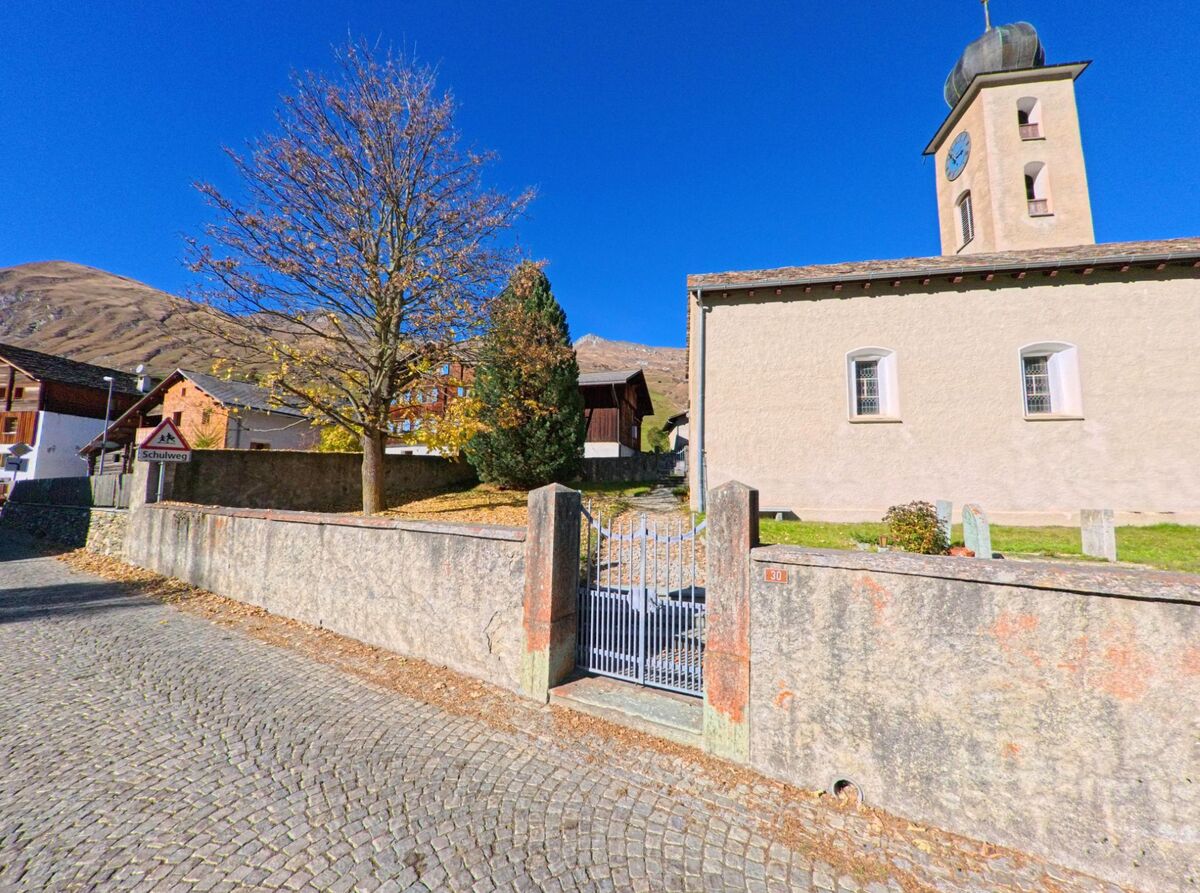 An einer gepflasterten Strasse in Rheinwald (7437, Kanton Graubünden) steht eine Kirche mit ummauertem Kirchhof, herbstlichen Bäumen und Berglandschaft unter klarem Himmel.