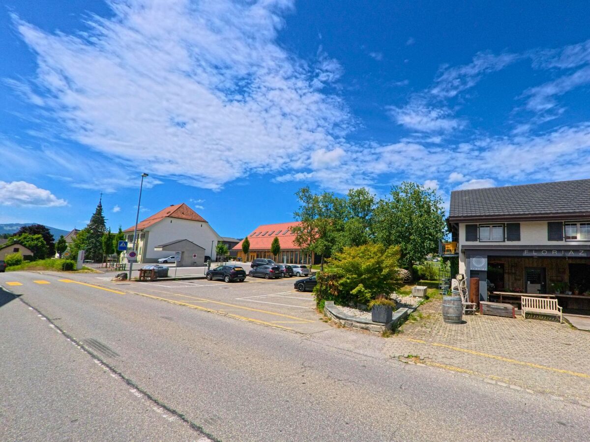 Entlang einer Hauptstrasse in Riaz (1632, Kanton Freiburg) befinden sich ein Blumenladen, ein Parkplatz und mehrere Gebäude mit roten Ziegeldächern.