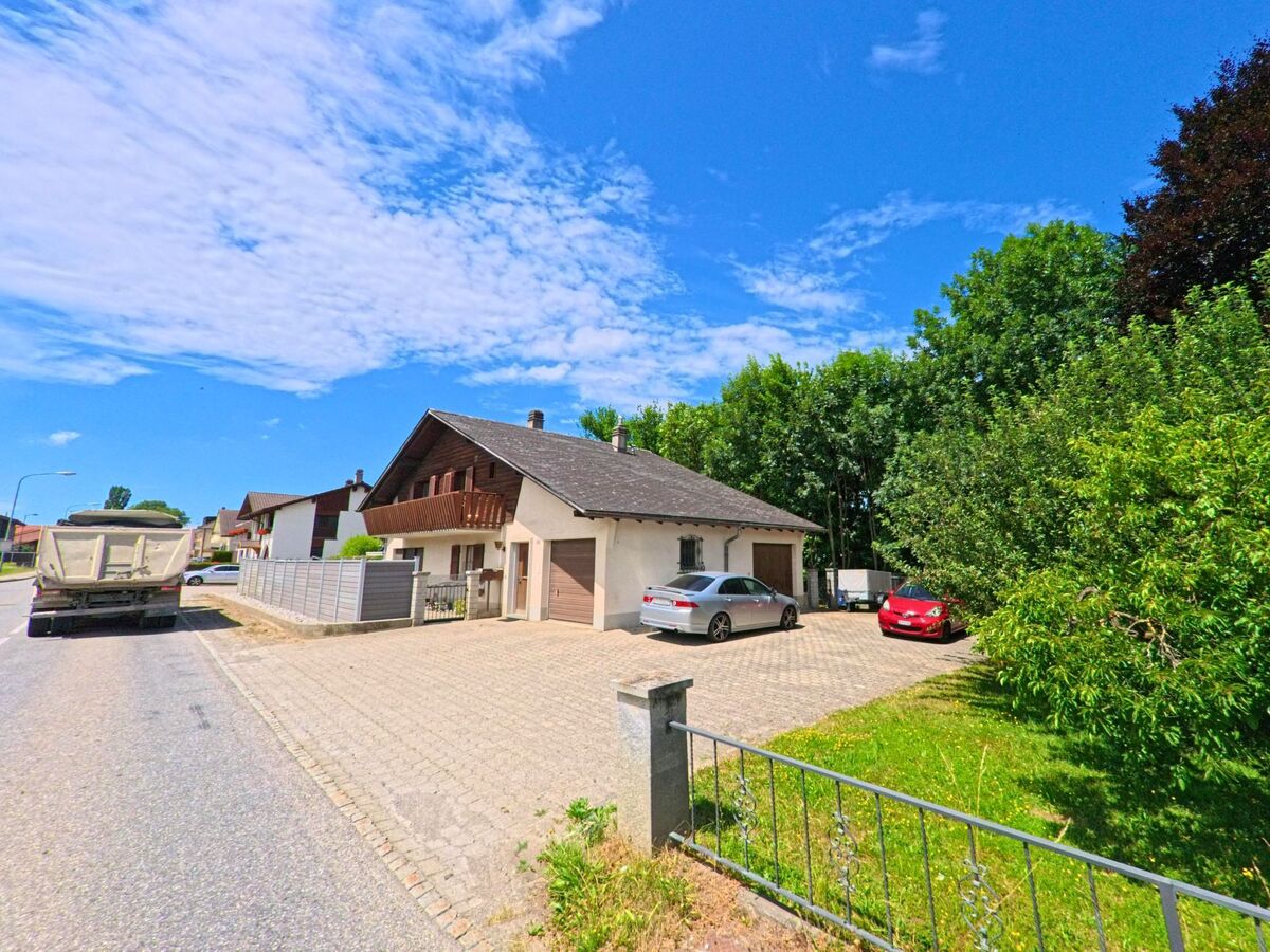 Ein Einfamilienhaus mit grossem Vorplatz und angrenzendem Garten steht in Riaz (1632, Kanton Freiburg) an einer ruhigen Strasse mit Sicht auf parkende Fahrzeuge und einen angrenzenden Baumstreifen.