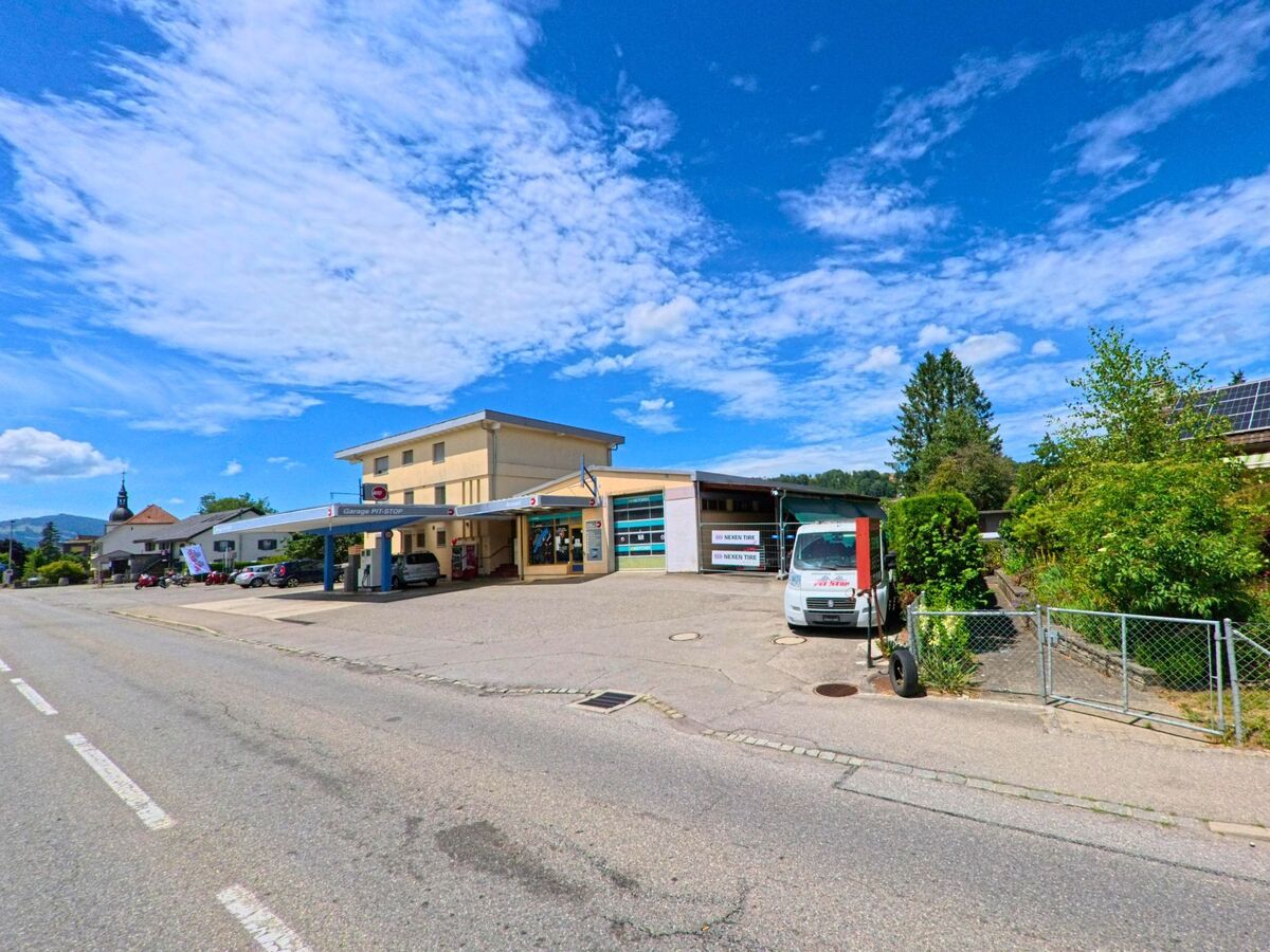 Eine Autogarage mit Tankstelle liegt in Riaz (1632, Kanton Freiburg) an einer breiten Strasse, im Hintergrund ist der Kirchturm des Dorfes zu sehen.