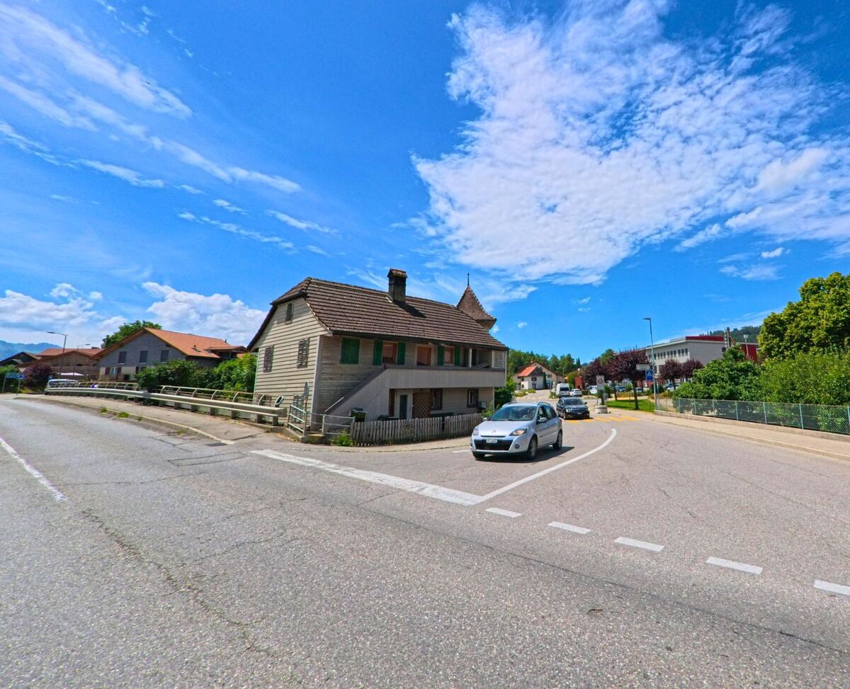 In Riaz (1632, Kanton Freiburg) verläuft eine verkehrsreiche Kreuzung an einem alten Wohnhaus mit grünen Fensterläden vorbei, flankiert von modernen Gebäuden im Hintergrund.